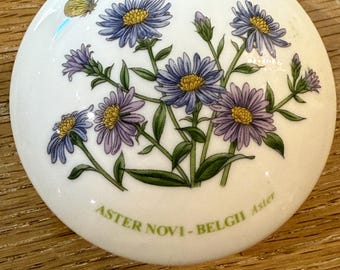 May include: A white, round ceramic decorative piece featuring a floral design with blue and purple aster flowers, green leaves, and a yellow butterfly. The word "September" is at the top, and "ASTER NOVI-BELGII Aster" is at the bottom.