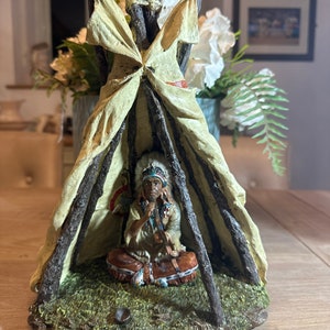 May include: A Native American figurine sitting inside a teepee made of brown sticks and a beige canvas. The figurine is wearing a feathered headdress and holding a pipe. The teepee is sitting on a green grassy base.