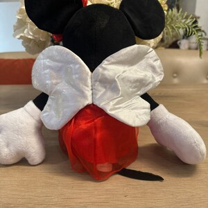 K&ouml;nnte beinhalten: Eine Pl&uuml;schpuppe von Minnie Mouse, von hinten gesehen, mit schwarzen Ohren, einer roten Schleife und wei&szlig;en Handschuhen. Die Puppe tr&auml;gt einen roten Rock mit einer wei&szlig;en, schimmernden Schleife. Die Puppe sitzt auf einem Holztisch.