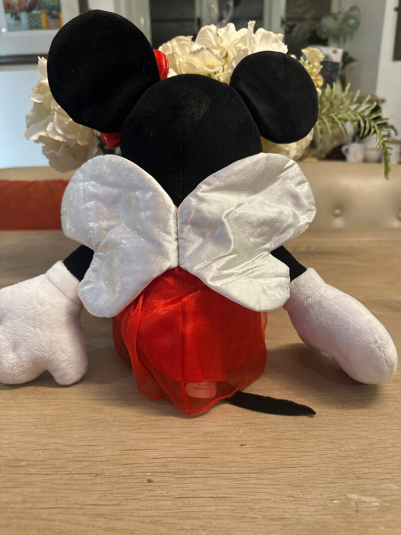 K&ouml;nnte beinhalten: Eine Pl&uuml;schpuppe von Minnie Mouse, von hinten gesehen, mit schwarzen Ohren, einer roten Schleife und wei&szlig;en Handschuhen. Die Puppe tr&auml;gt einen roten Rock mit einer wei&szlig;en, schimmernden Schleife. Die Puppe sitzt auf einem Holztisch.