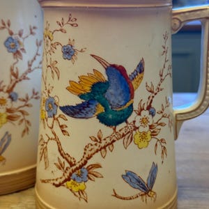 May include: Cream-colored ceramic pitcher with a detailed bird and floral design. The bird is depicted with blue, red, and yellow feathers, perched on a branch with blue and yellow flowers. The pitcher has a tan base and handle.
