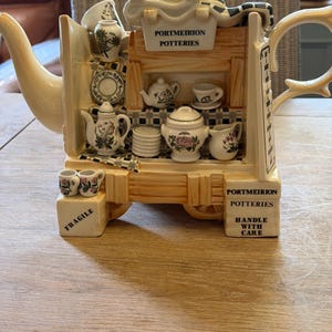 Può includere: Una teiera in ceramica color crema a forma di vetrina, con set da tè in miniatura, piattini e motivi floreali. La struttura presenta insegne con le scritte PORTMEIRION POTTERIES, FRAGILE e HANDLE WITH CARE e un tetto a scacchi bianco e nero.