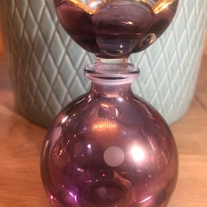 Op de afbeelding: Een paarse glazen fles met witte stippen en een gouden bloemvormige dop.