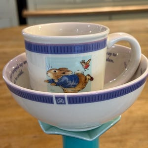 Op de afbeelding: Een witte keramische kom en mok set met een Peter Rabbit ontwerp. De mok zit in de kom. De kom en mok hebben blauwe en paarse randen. De mok heeft een oor. De kom staat op een turquoise standaard.