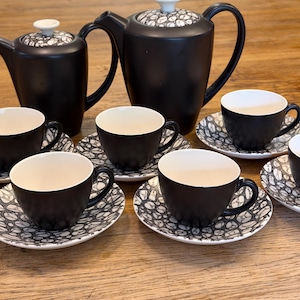 May include: A tea set featuring two matte black teapots and several matching teacups with saucers. Each piece has a white interior and a lid or saucer decorated with a black and white pebble-like pattern. The teapots have white knobs on the lids.