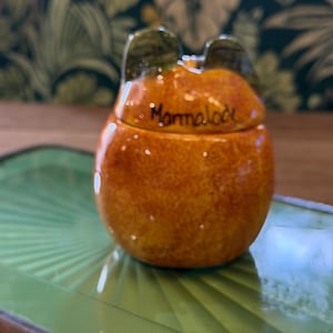 May include: A round, orange ceramic jar with a lid featuring two small, dark green ears. The word Marmalade is written in black cursive script on the front of the lid. The jar has a mottled, textured finish and a glossy, reflective surface.
