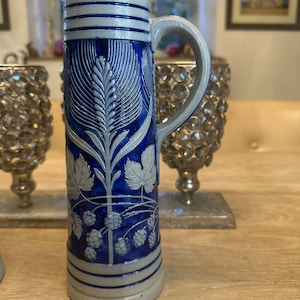 Op de afbeelding: Een blauwe en witte keramische bierkan met een handvat. De kan heeft een ontwerp van tarwehalmen en hop.