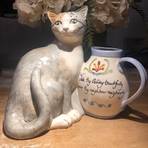 May include: A ceramic cat figurine with grey, white and orange fur sits next to a blue and white ceramic pitcher with the text "Take thy Calling thankfully love thy neighbour neighbourly".