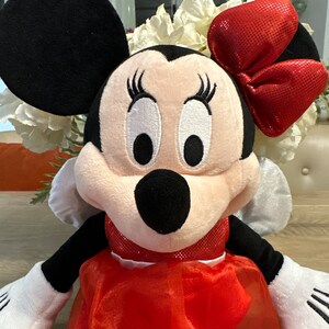 K&ouml;nnte beinhalten: Eine Minnie Mouse Pl&uuml;schpuppe mit schwarzem Kopf und Ohren, einer roten Schleife und einem roten Kleid. Die Puppe hat wei&szlig;e Handschuhe und ein l&auml;chelndes Gesicht mit gro&szlig;en Augen und Wimpern. Im Hintergrund sind wei&szlig;e Blumen zu sehen.