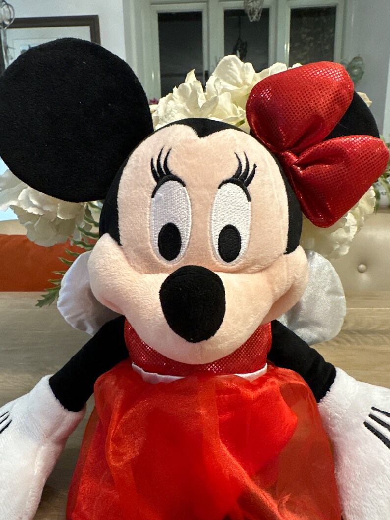 K&ouml;nnte beinhalten: Eine Minnie Mouse Pl&uuml;schpuppe mit schwarzem Kopf und Ohren, einer roten Schleife und einem roten Kleid. Die Puppe hat wei&szlig;e Handschuhe und ein l&auml;chelndes Gesicht mit gro&szlig;en Augen und Wimpern. Im Hintergrund sind wei&szlig;e Blumen zu sehen.