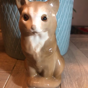 May include: A ceramic cat figurine in a seated position. The cat has a tan and white coat with brown eyes. The figurine is glossy and has a detailed design, showcasing a classic cat pose. The cat is a decorative item.