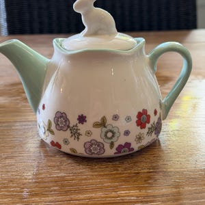 May include: A white ceramic teapot featuring a light green handle and spout, decorated with a colourful floral pattern of red, purple, and blue flowers. The lid is topped with a small, white, sculpted rabbit figurine as a decorative handle.