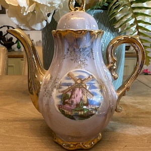 May include: A vintage porcelain teapot with a delicate floral design and a gold trim. The teapot features a windmill scene with a pink roof and blue sky. The teapot has a gold handle and spout.
