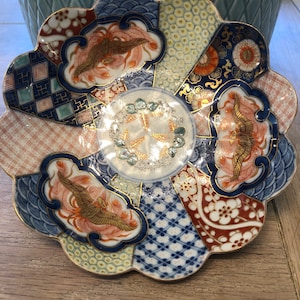 Puede incluir: Un cuenco de cerámica decorativo con un borde festoneado. El cuenco está decorado con un diseño colorido de inspiración japonesa que presenta detalles dorados, patrones geométricos azules y blancos y pájaros rojos y dorados.
