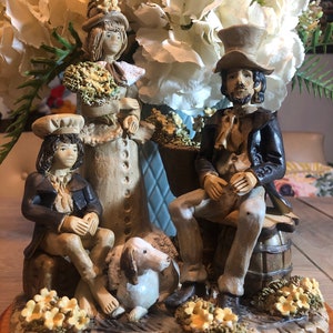 May include: A ceramic figurine set of a family with a dog. The family is dressed in vintage clothing and the father is wearing a top hat. The figurine is sitting on a wooden base with a floral design.