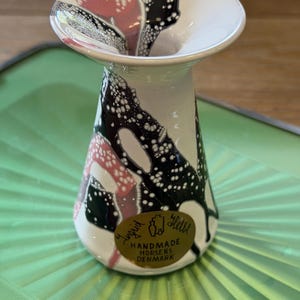 Puede incluir: Un pequeño jarrón de cerámica con borde acampanado, que presenta una base blanca decorada con patrones abstractos moteados en negro, blanco y rosa. Una pegatina dorada ovalada en el frente dice Ingrid Helle Handmade Horsens Denmark con un icono de botella.