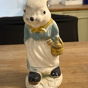 May include: A vintage ceramic rabbit figurine, painted in off-white, blue, and yellow. The rabbit wears a blue shirt, white apron, and black shoes, holding a small basket. The figurine has a charming, aged appearance, with visible wear and tear.