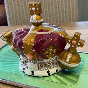 Può includere: Una teiera in ceramica a forma di corona reale, caratterizzata da una parte superiore color bordeaux intenso con texture vellutata, una fascia dorata con accenti bianchi e verdi e una base bianca in stile ermellino. Include un manico a forma di globo.