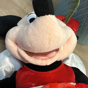 K&ouml;nnte beinhalten: Eine Pl&uuml;schpuppe von Minnie Mouse mit einem roten Paillettenkleid und einer Schleife. Die Puppe hat schwarze Ohren, ein rosa Gesicht und wei&szlig;e Fl&uuml;gel. Die Puppe liegt auf einer Holzoberfl&auml;che.
