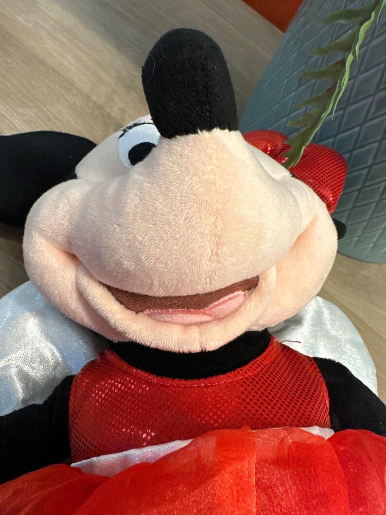 K&ouml;nnte beinhalten: Eine Pl&uuml;schpuppe von Minnie Mouse mit einem roten Paillettenkleid und einer Schleife. Die Puppe hat schwarze Ohren, ein rosa Gesicht und wei&szlig;e Fl&uuml;gel. Die Puppe liegt auf einer Holzoberfl&auml;che.