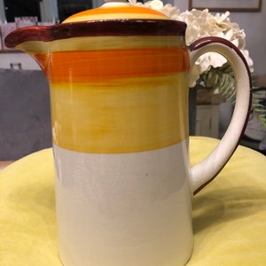 Puede incluir: Una cafetera de cerámica con un diseño de rayas marrones, naranjas y amarillas. La cafetera tiene una tapa con un pomo en la parte superior.