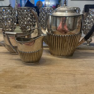 May include: A silver teapot and creamer set with a decorative, fluted design. The teapot has a lid with a finial and a spout. The creamer has a handle and a spout. The set is on a wooden surface.