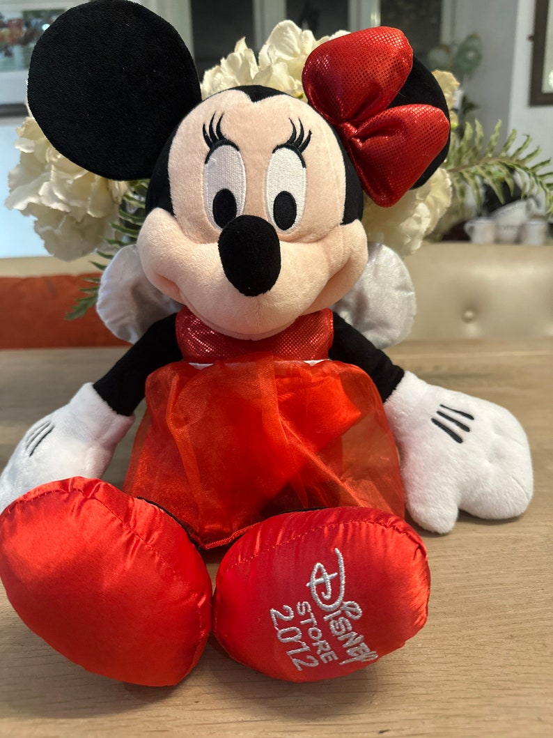 K&ouml;nnte beinhalten: Minnie Mouse Pl&uuml;schtier mit roter Schleife, Kleid und Schuhen. Das schwarz-wei&szlig;e Pl&uuml;schtier hat wei&szlig;e Handschuhe und schwarze Ohren. Das Logo Disney Store 2012 ist auf einem der roten Schuhe gestickt.