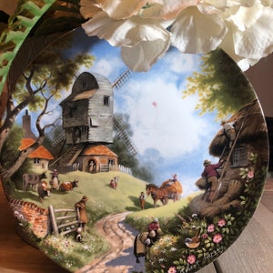 Puede incluir: Un plato de porcelana vintage con una pintura colorida de un molino de viento, un camino de campo y una cabaña con techo de paja. El plato está decorado con flores y tiene la firma del artista, "Robert Hersey", en la esquina inferior derecha.