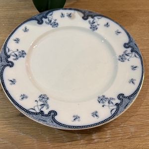 Puede incluir: Plato de cerámica blanca con un diseño floral y de volutas azules alrededor del borde. El plato tiene un borde dorado y una estética simple y elegante. El plato es probablemente para servir comida.