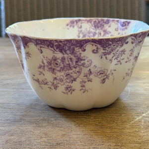 May include: A white ceramic bowl featuring a scalloped rim and a decorative purple floral transferware pattern. The design consists of intricate blossoms and scrolling foliage that wraps around the upper exterior of the rounded vessel.