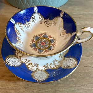Puede incluir: Un juego de taza y platillo de té vintage con un diseño floral azul y blanco. La taza y el platillo tienen detalles dorados y un delicado patrón de encaje.