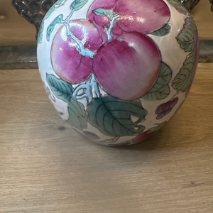 Può includere: Un vaso in ceramica bianca con un design floreale che presenta frutta rosa e verde. Il vaso ha una forma rotonda e una finitura liscia e lucida.