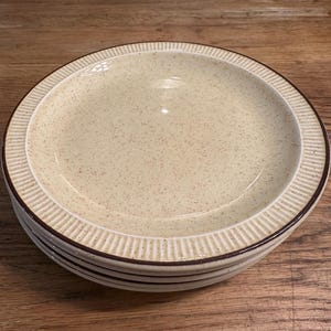 May include: A stack of four beige ceramic dinner plates with a brown rim and a speckled texture.