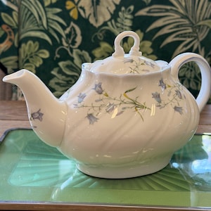 Puede incluir: Una tetera de porcelana blanca con un delicado patrón floral de flores azules en forma de campana y tallos verdes. La tetera tiene un asa curva, una tapa festoneada con un asa de bucle y un cuerpo acanalado con un diseño en espiral.