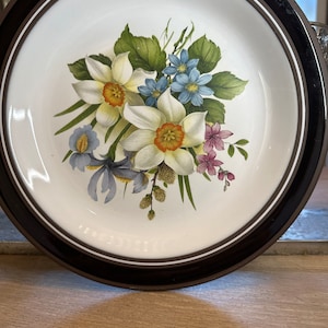 Könnte beinhalten: Ein weißer Keramikteller mit braunem Rand zeigt ein Blumenmuster aus weißen, gelben, blauen und rosa Blüten. Die Blumen sind in einem Strauß angeordnet und von grünen Blättern umgeben.