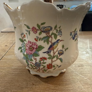May include: A white ceramic planter with a scalloped rim and a floral design featuring a blue bird, pink, red, and yellow flowers, and green leaves.