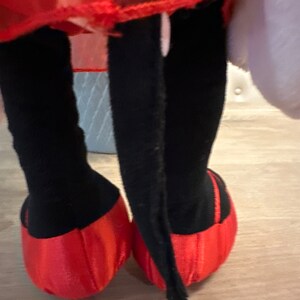 K&ouml;nnte beinhalten: Ein Pl&uuml;schtier mit schwarzen Beinen und roten Schuhen. Das Spielzeug hat einen roten Rock und ein wei&szlig;es, flauschiges Element. Die Schuhe sind leuchtend rot und die Beine sind schwarz.