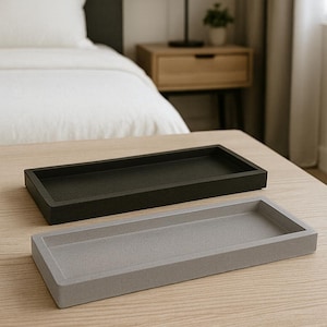 May include: Two rectangular trays, one dark gray and one light gray, sit side-by-side on a light wood surface. The trays have a simple, minimalist design with raised edges. The background includes a bed, nightstand, and lamp.