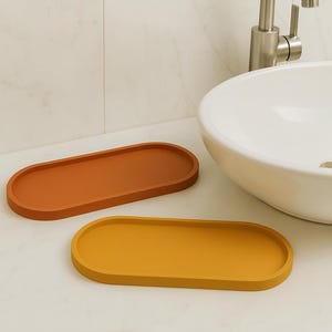 May include: Two oval-shaped trays in the colors of orange and yellow. The trays are made of a solid material and have a smooth surface. The trays are placed on a white countertop near a white sink and a silver faucet.