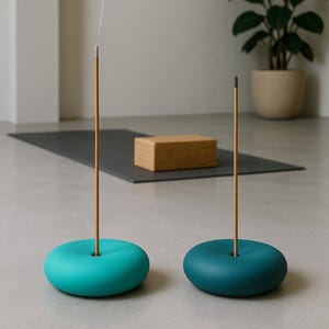 May include: Two teal incense holders with incense sticks, one emitting smoke. A grey yoga mat and a wooden block are in the background. A potted plant is also visible. The holders are round and have a hole in the centre for the incense.