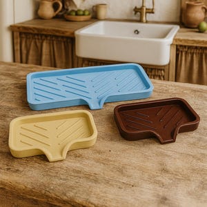 May include: Three soap dishes in blue, yellow, and brown sit on a wooden surface. The rectangular dishes have a unique shape with a sloped design and a small spout. The background features a kitchen sink and decorative items.