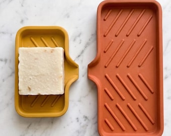 May include: A rectangular mustard yellow soap dish holding a square off-white bar of soap and a larger rectangular terracotta orange soap dish with a drainage spout and diagonal raised ridges on the interior surface of both trays.
