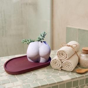 May include: A light purple, torso-shaped planter containing a small green succulent, resting on a dark maroon oval tray. Beside it are three rolled beige towels tied with twine, a glass jar filled with white bath salts, and a small wooden spoon.