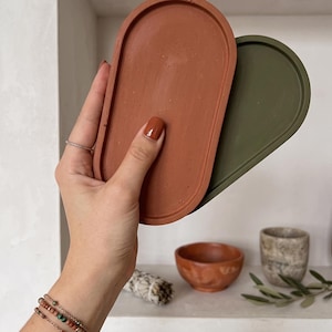 May include: Two oval-shaped trays, one terracotta and one olive green, are held in a hand. The trays are made of a matte material. Other items on a shelf include a small terracotta bowl, a speckled ceramic cup, and a bundle of sage.