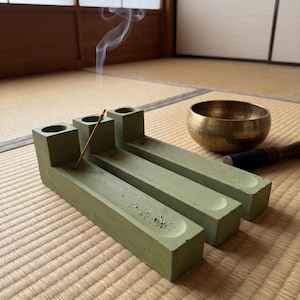May include: A sage green concrete incense holder with three cylindrical openings and long rectangular channels, featuring a single burning incense stick emitting a thin trail of smoke, positioned next to a metallic gold singing bowl.