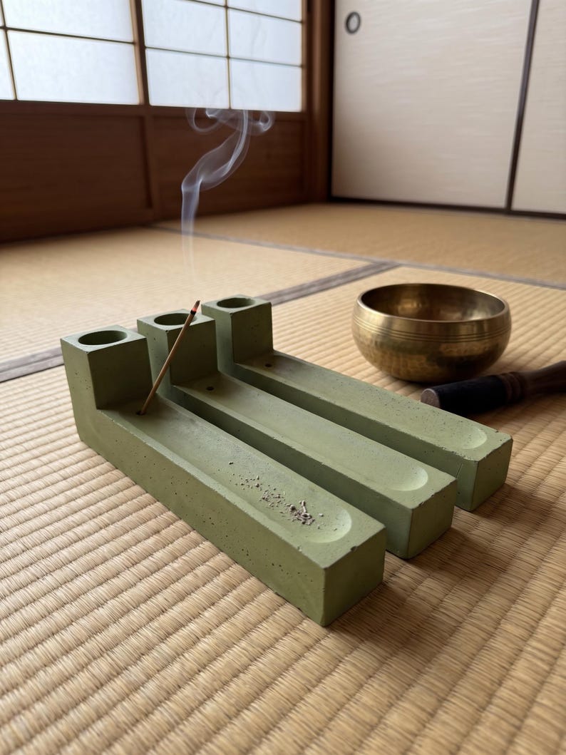 May include: A sage green concrete incense holder with three cylindrical openings and long rectangular channels, featuring a single burning incense stick emitting a thin trail of smoke, positioned next to a metallic gold singing bowl.