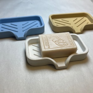 May include: Three soap dishes made of concrete in light blue, beige, and white. The white dish has a bar of soap on top. The soap is white and has the word "Luxury" printed on it.