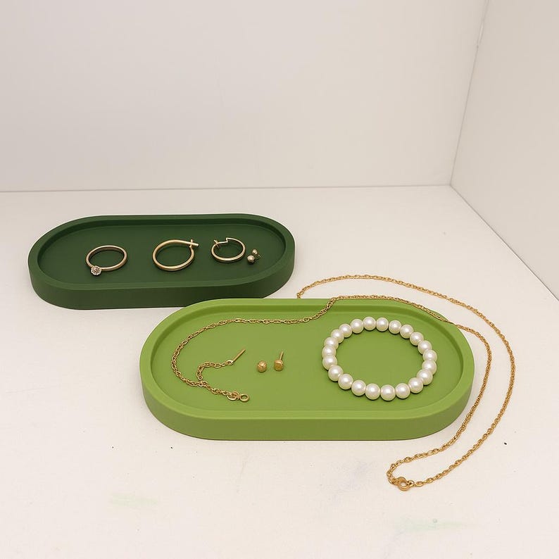 May include: Two oval-shaped jewelry trays in green. The top tray is a darker green and holds gold rings and earrings. The bottom tray is a lighter green and holds a pearl bracelet, gold necklace, and earrings.