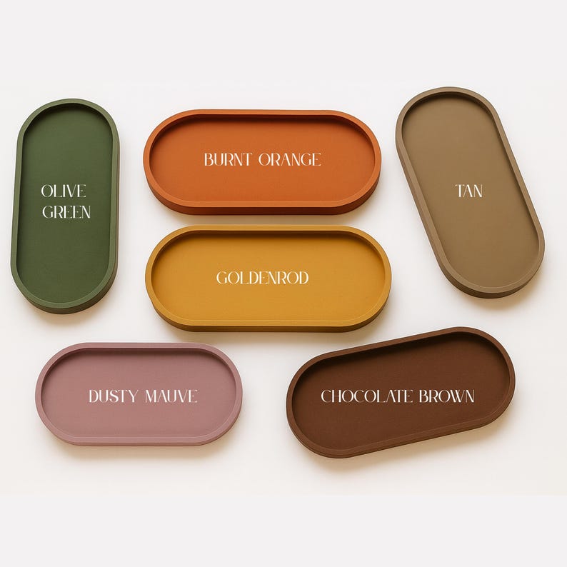 May include: A collection of six oval-shaped trays in various colors: olive green, burnt orange, goldenrod, tan, dusty mauve, and chocolate brown. Each tray has a slightly raised edge and a matte finish, with the color name printed on each.