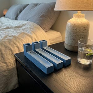 May include: Three rectangular blue concrete incense holders arranged side by side. Each holder features a cylindrical opening at one end and a long, narrow recessed channel extending along the top surface for holding incense sticks.